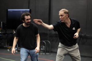 Two men interact in a studio; one gestures with his hand while the other stands nearby, watching. Electronic equipment and cables are visible in the background.