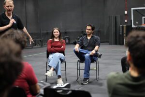 Two young adults sit on chairs facing a group, listening while a standing man gestures and speaks in a large indoor space.