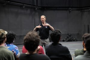 A man stands in front of a small seated audience, gesturing with his hands while speaking in a dark indoor setting.