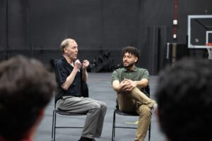 Two men sit on chairs having a conversation in a gymnasium; one gestures with his hands while the other listens with legs crossed.