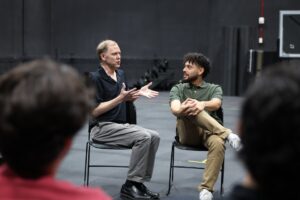 Two men sit and talk in chairs facing an audience in a large indoor space with black walls, while two people listen in the foreground.