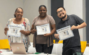 Three people stand indoors holding "APA World Games Silver Medal Winner" certificates and smiling at the camera. A laptop and papers are on the table in front of them.