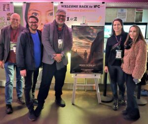 Five people stand indoors in front of film posters and a sign that reads "WELCOME BACK TO IFC Theater Entrances." A movie poster is displayed on an easel in the center.