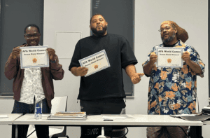 Three men stand behind a table, each holding a certificate that reads "APA World Games Bronze Medal Winner." They are posing for a photo indoors.