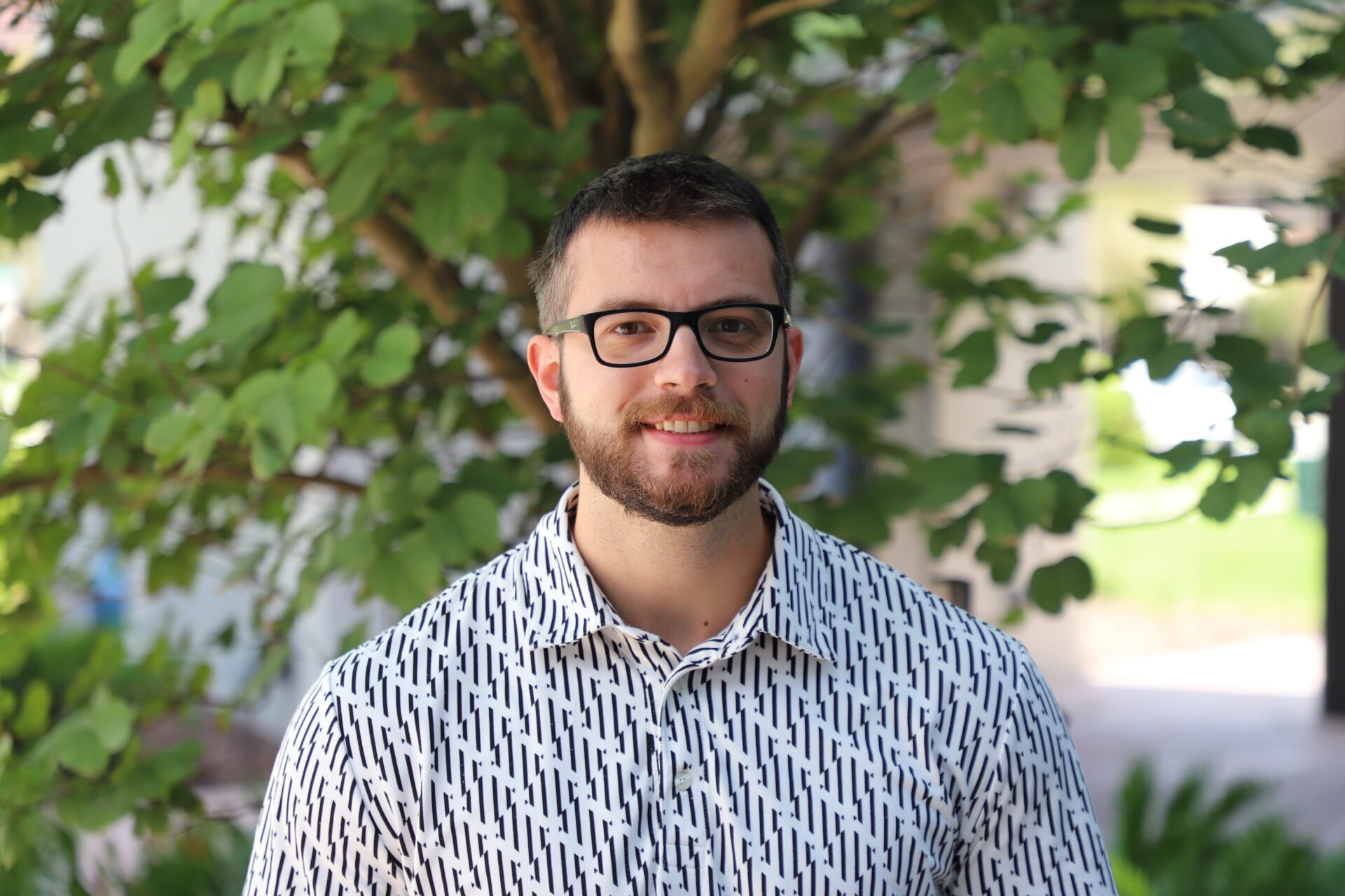 A man with glasses and a beard, wearing a white patterned shirt, stands outdoors in front of leafy green branches.