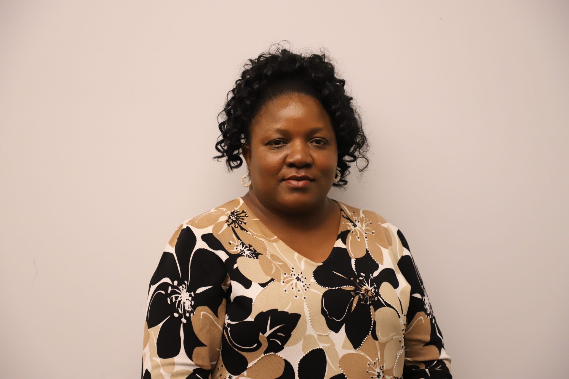 Woman with curly hair stands against a plain, light-colored wall, wearing a beige and black floral-patterned top.