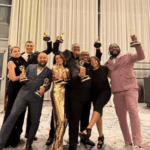A group of eight people dressed in formal attire pose indoors, each holding up an Emmy award and smiling at the camera.