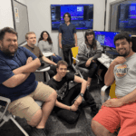 Seven people are gathered in an office with computers and monitors, some seated and some standing, posing and smiling for the camera.
