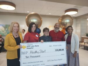 Five people stand indoors, holding a large $10,000 check from UCF Lake Nona Hospital to Healthy Start. Festive balloons are in the background.