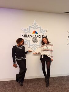 Two women stand smiling in front of a Milan Cortina 2026 Winter Olympics sign, each gesturing toward the logo on the wall.