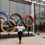 A person stands in front of the Olympic rings and NBC Sports building, posing with hands on hips and smiling at the camera.