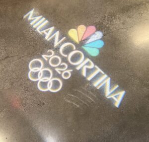 Logo projection on a concrete floor showing the NBC peacock and text: "Milan Cortina 2026" with Olympic rings, promoting the 2026 Winter Olympic Games.