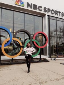 A person stands in front of the Olympic rings and NBC Sports building, posing with hands on hips and smiling at the camera.