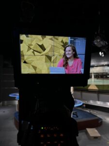 A woman in a pink shirt sits at a news desk, visible on a camera monitor in a television studio with geometric yellow background graphics.