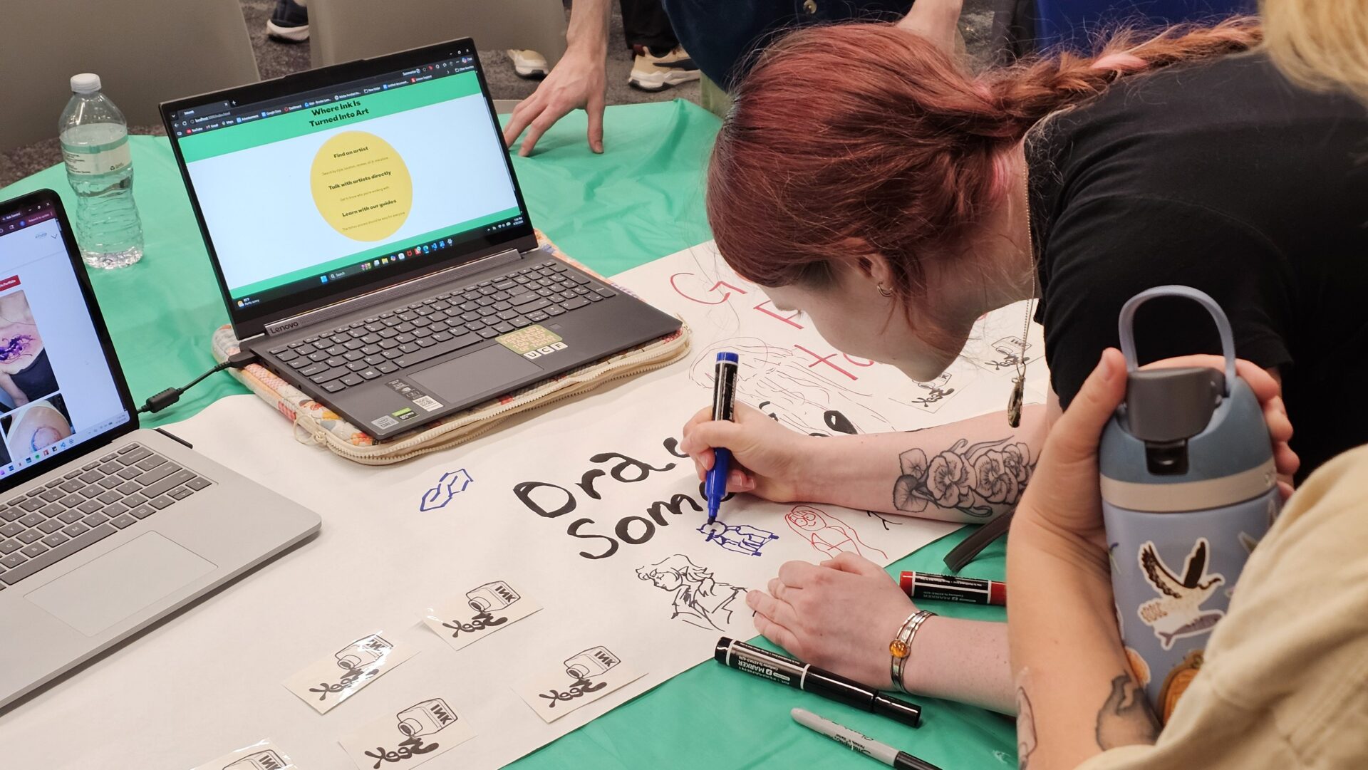 A person with red hair draws on a large sheet of paper beside two open laptops, while another person holds a water bottle and several markers are on the table.
