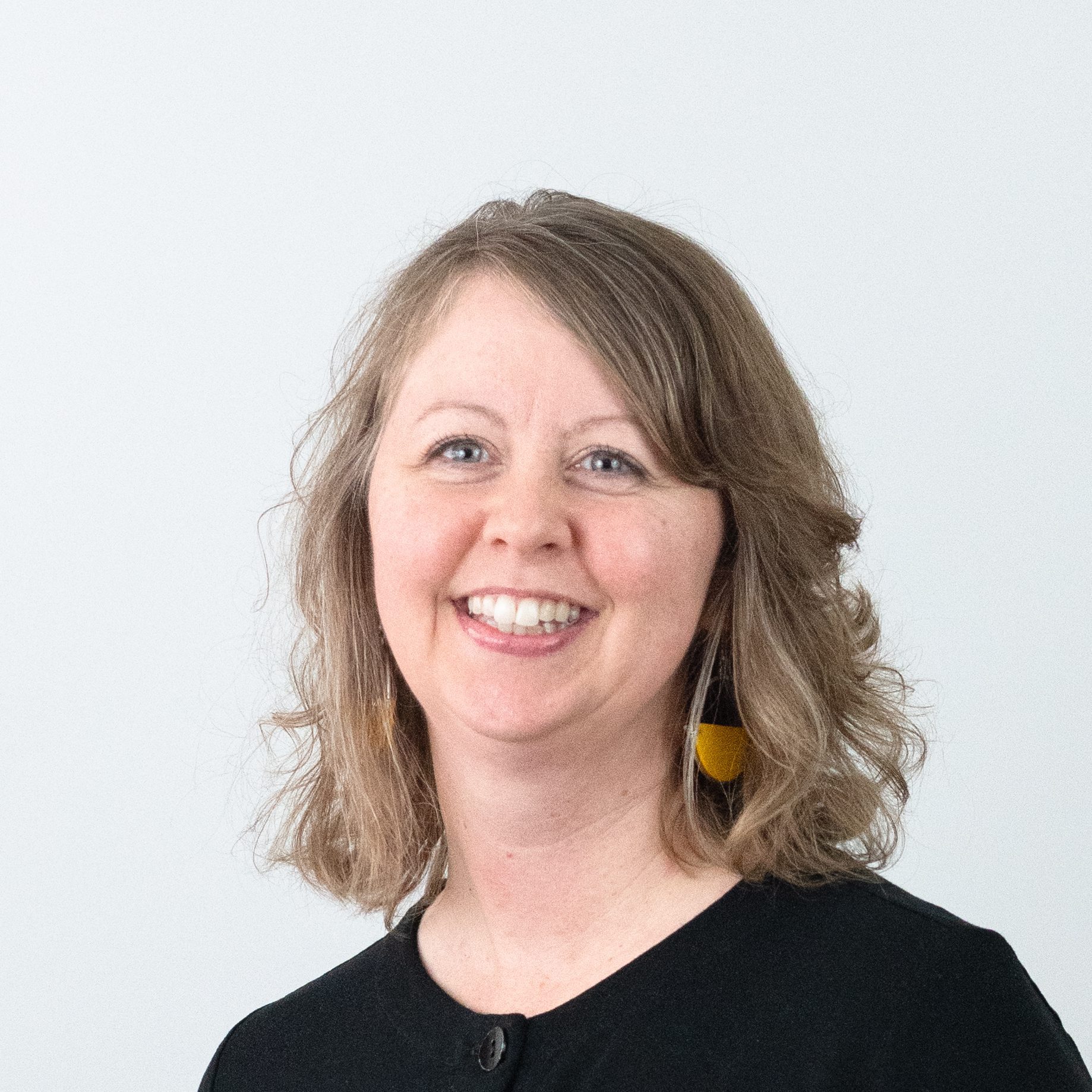Smiling woman with wavy, shoulder-length light brown hair wearing a black top and yellow earrings, standing in front of a plain white background.