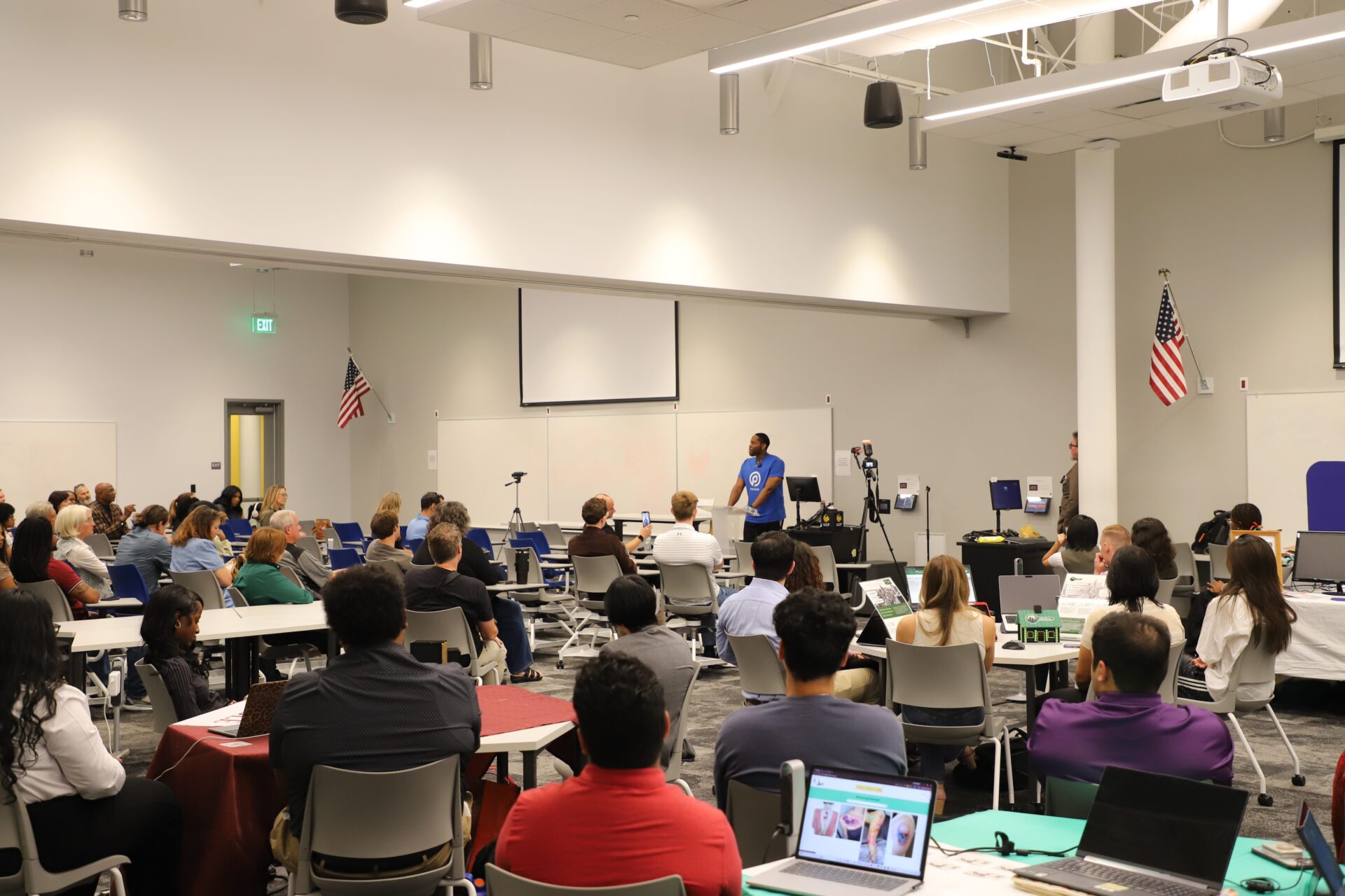A speaker stands at the front of a conference room addressing an audience seated at tables, with laptops open and American flags displayed on the walls.
