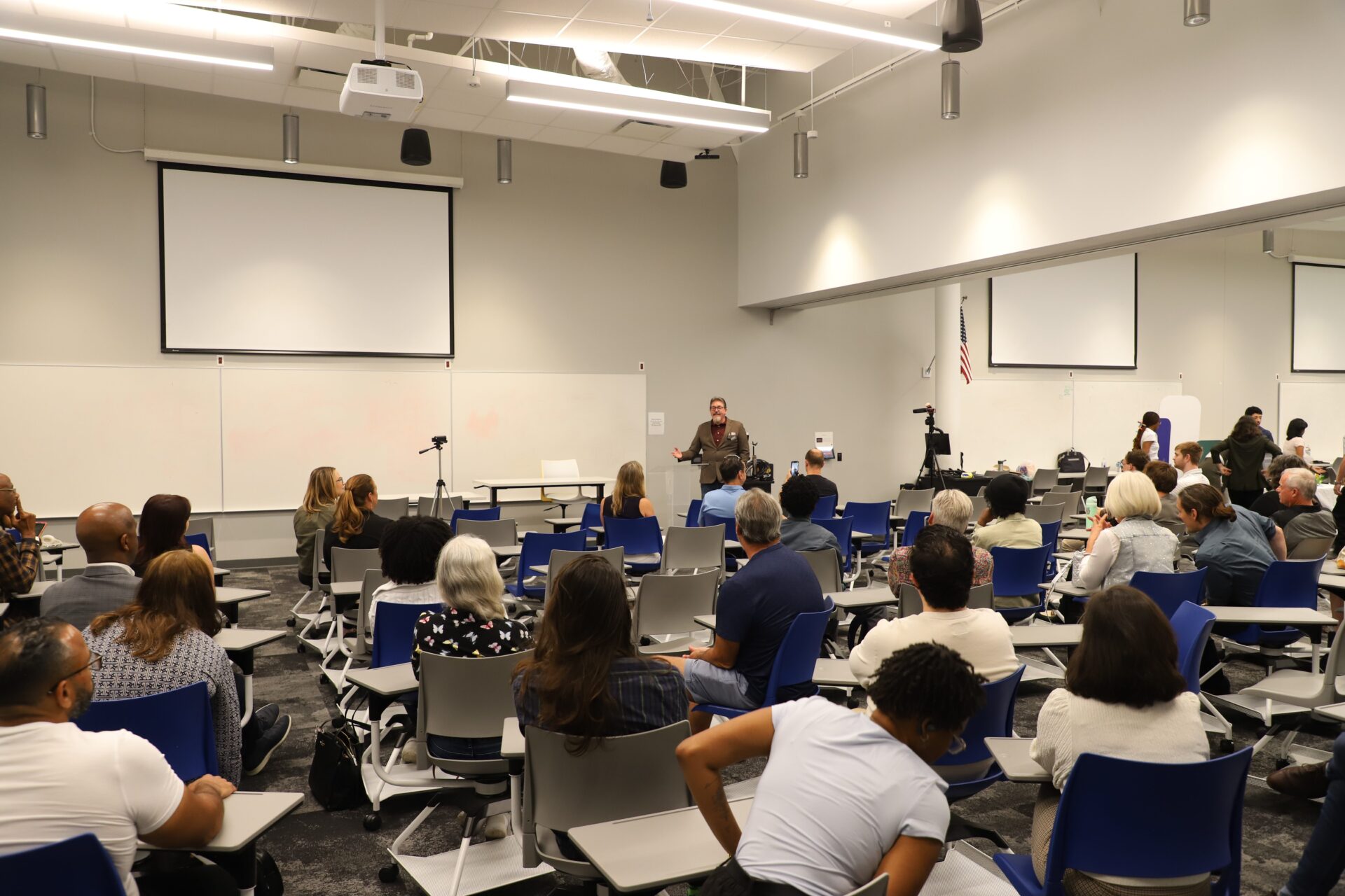 A group of people sits in a modern classroom with blue chairs, facing a speaker at the front near a whiteboard and projection screens.