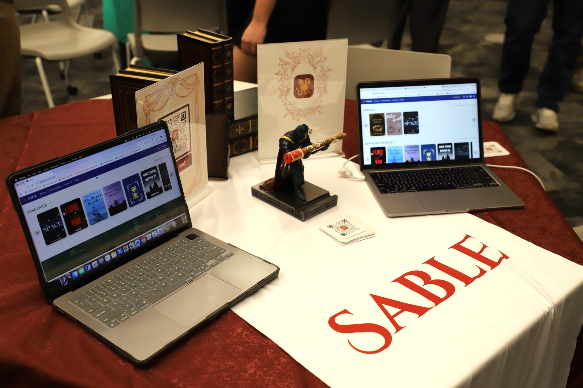 Two open laptops sit on a table with books, a figurine, and a sign labeled "SABLE." Both laptops display book-related content on their screens.