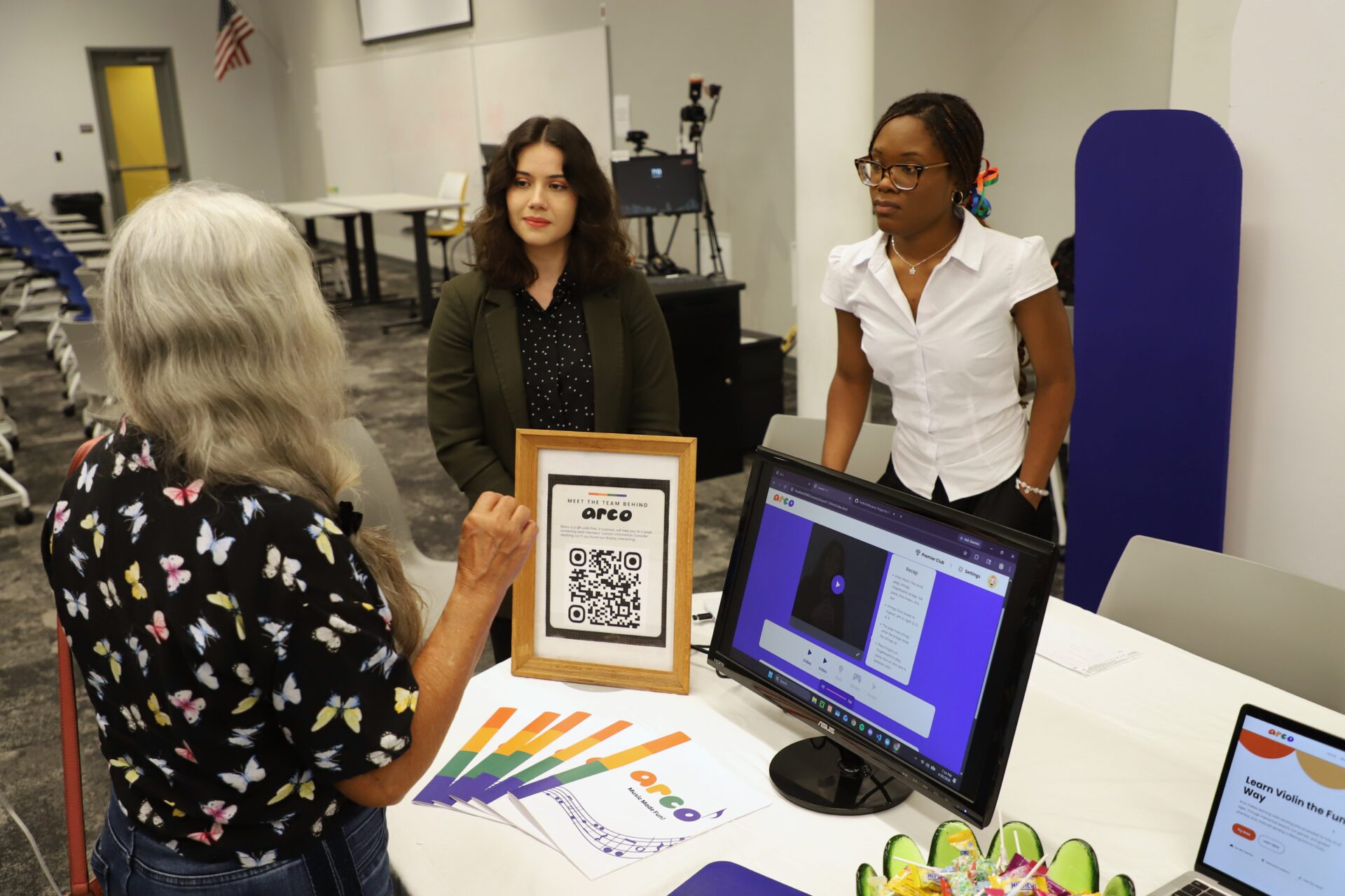 Three women stand around a table at a booth with informational materials, a framed QR code, a computer monitor, and brochures in a conference room setting.