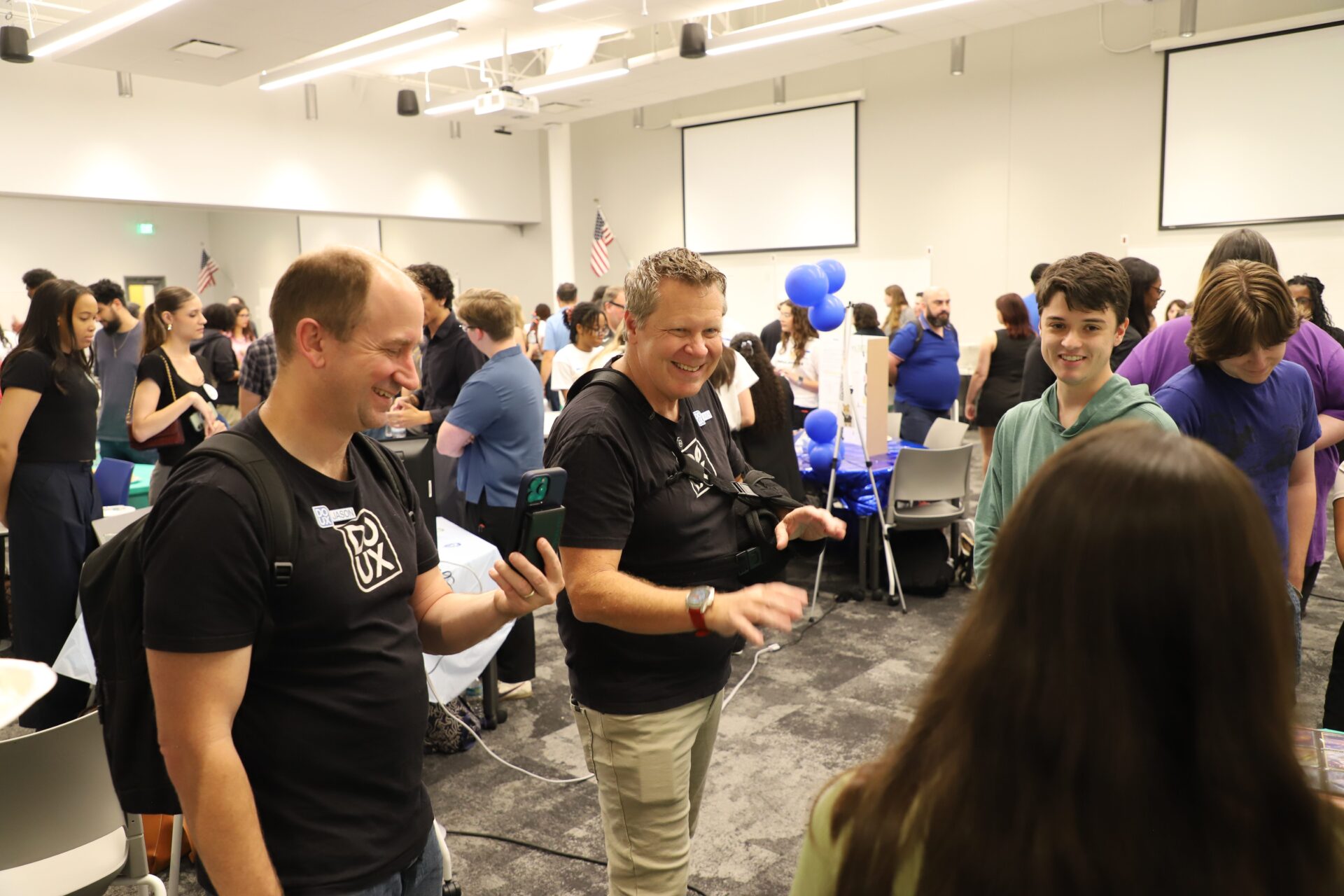 People interact and smile at a busy indoor event with tables, balloons, and informational displays in the background.