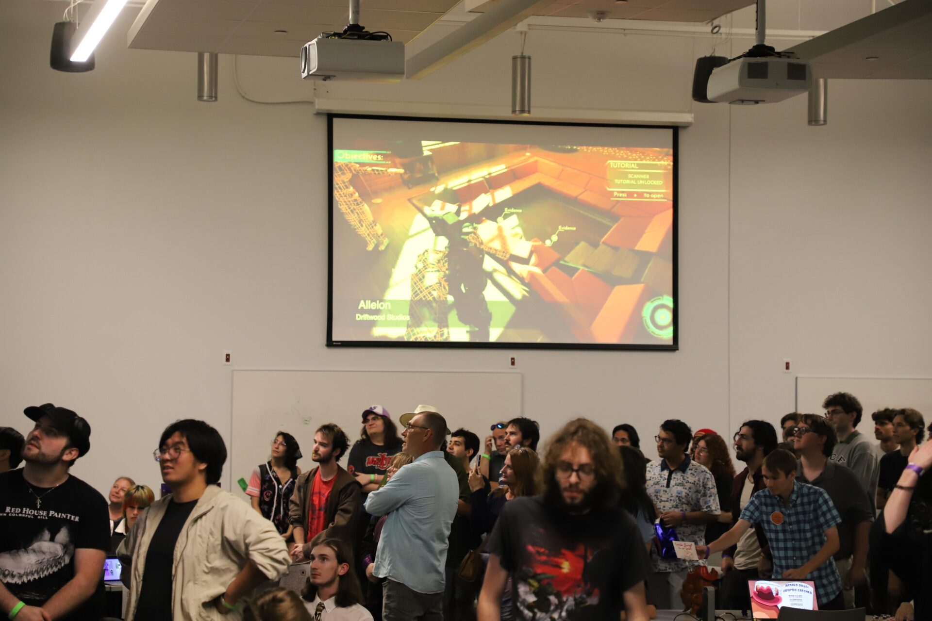 A group of people watch a large screen displaying a video game in a brightly lit room with white walls and ceiling-mounted projectors.