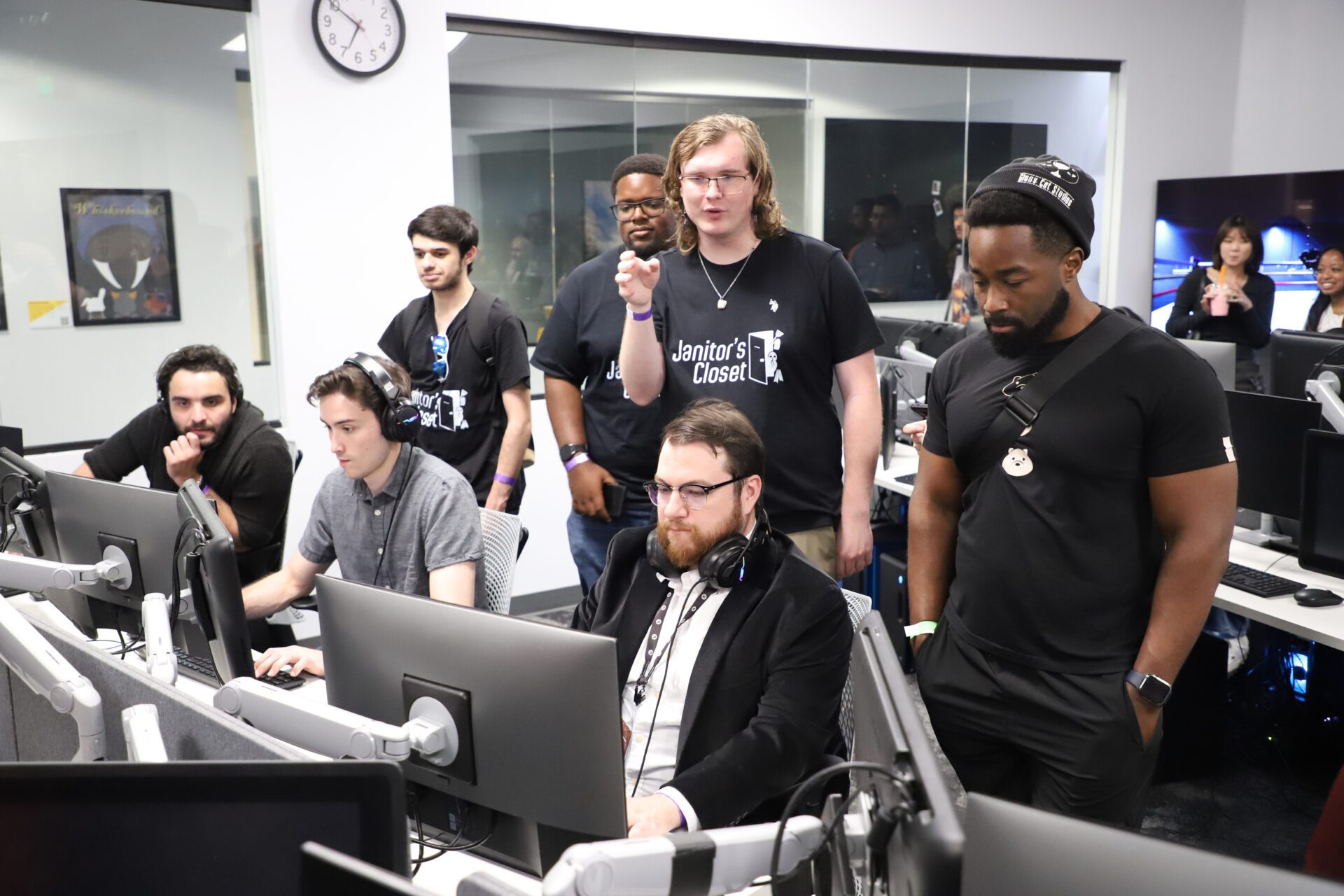 A group of people in a computer lab, some seated and working at monitors while others stand behind them observing or discussing.