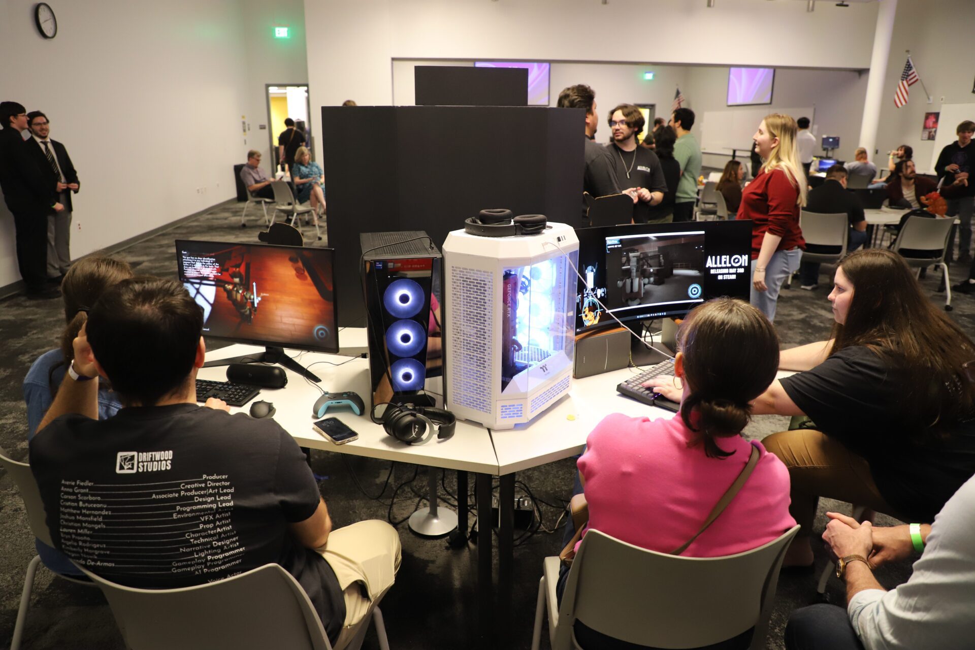 A group of people sit around a table with gaming computers and monitors at an indoor event, while others stand and watch in the background.