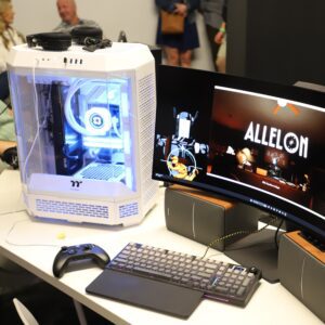 A white gaming PC with LED lights is on a desk next to a curved monitor displaying the game ALLELION, along with a keyboard, mouse, controller, and speakers.