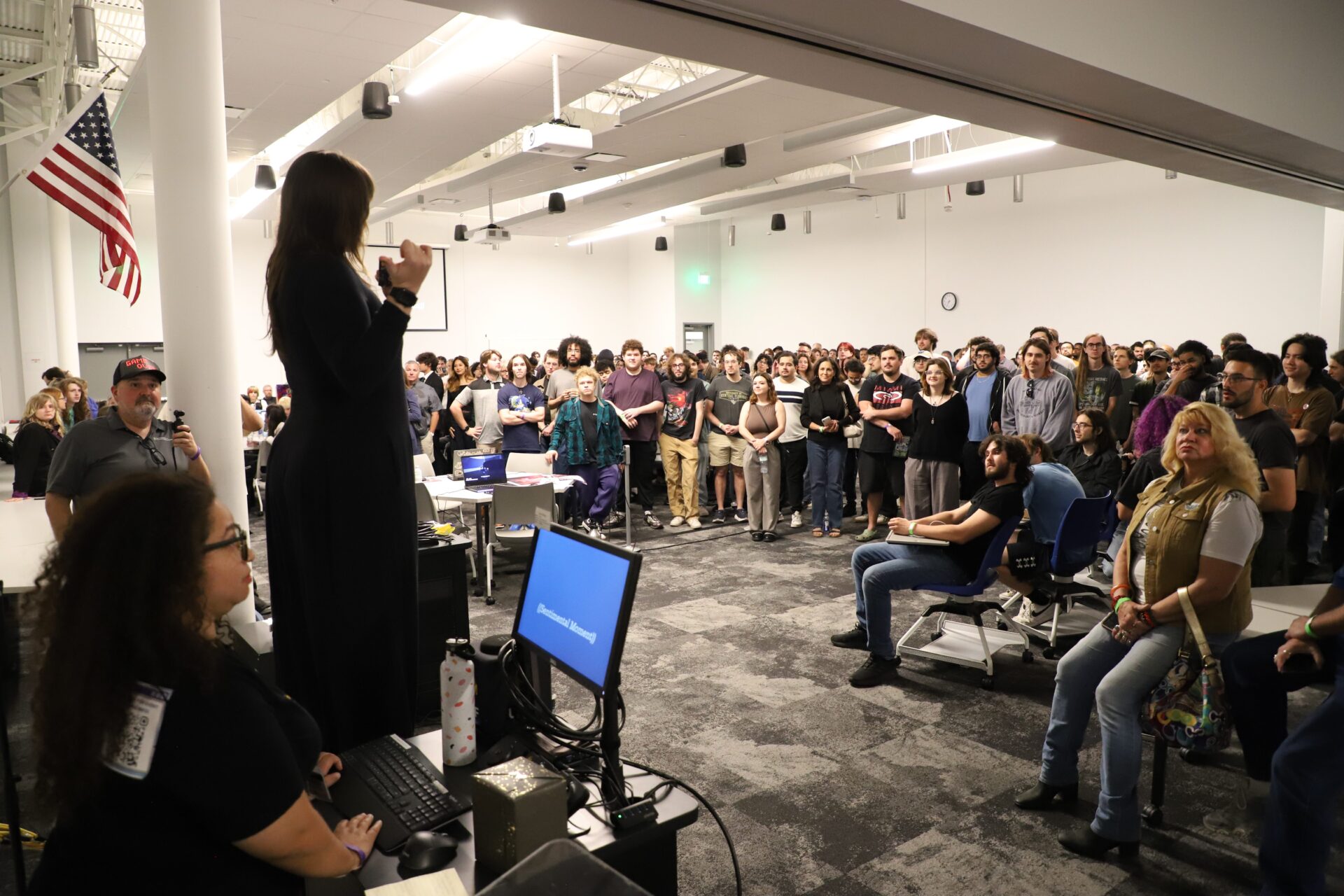 A woman stands and speaks to a large group of people gathered in a modern classroom or meeting space, with an American flag visible on the left.