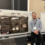 A man stands and smiles next to a presentation board titled "Chocolate Kingdom" at a conference or exhibition. The board displays sections on the problem, solution, and future work.