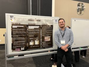 A man stands and smiles next to a presentation board titled "Chocolate Kingdom" at a conference or exhibition. The board displays sections on the problem, solution, and future work.