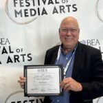 A man in glasses and a suit holds a certificate in front of a "Festival of Media Arts" backdrop.