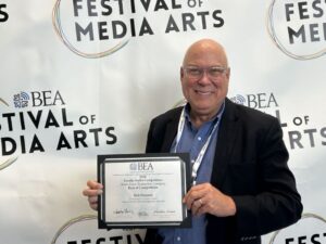A man in glasses and a suit holds a certificate in front of a "Festival of Media Arts" backdrop.