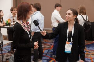 A woman holding a microphone interviews another woman at a conference; several people are conversing in the background.