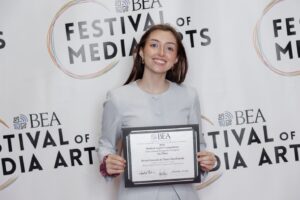 A woman stands smiling and holding a certificate in front of a backdrop that reads "BEA Festival of Media Arts.