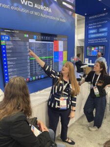 A woman points at a large display screen showing a software interface while two others observe at a trade show booth.