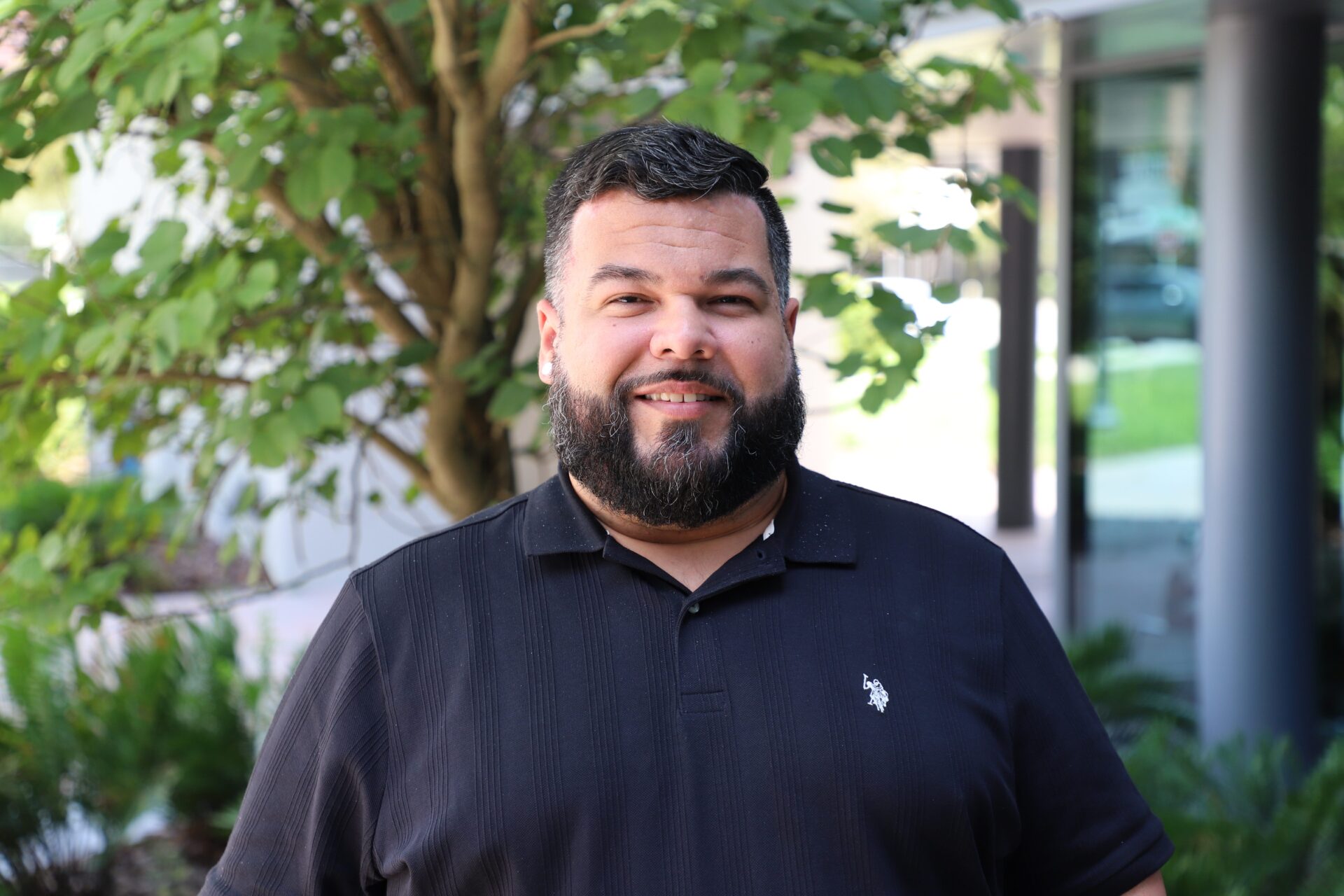 A man with a beard and short hair stands outdoors in front of a tree, wearing a black collared shirt and smiling at the camera.