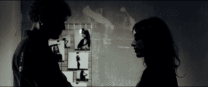 Two people stand facing each other in silhouette; behind them is a wall with several black-and-white photos of a woman in various poses.