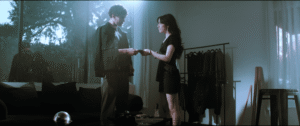 A man and woman stand in a dimly lit room, facing each other and exchanging an envelope, with clothes hanging on a rack and large windows in the background.