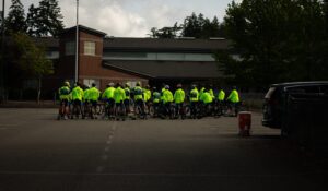 A group of cyclists in bright yellow jackets gather in a parking lot near a brick building, with trees and parked cars in the background.