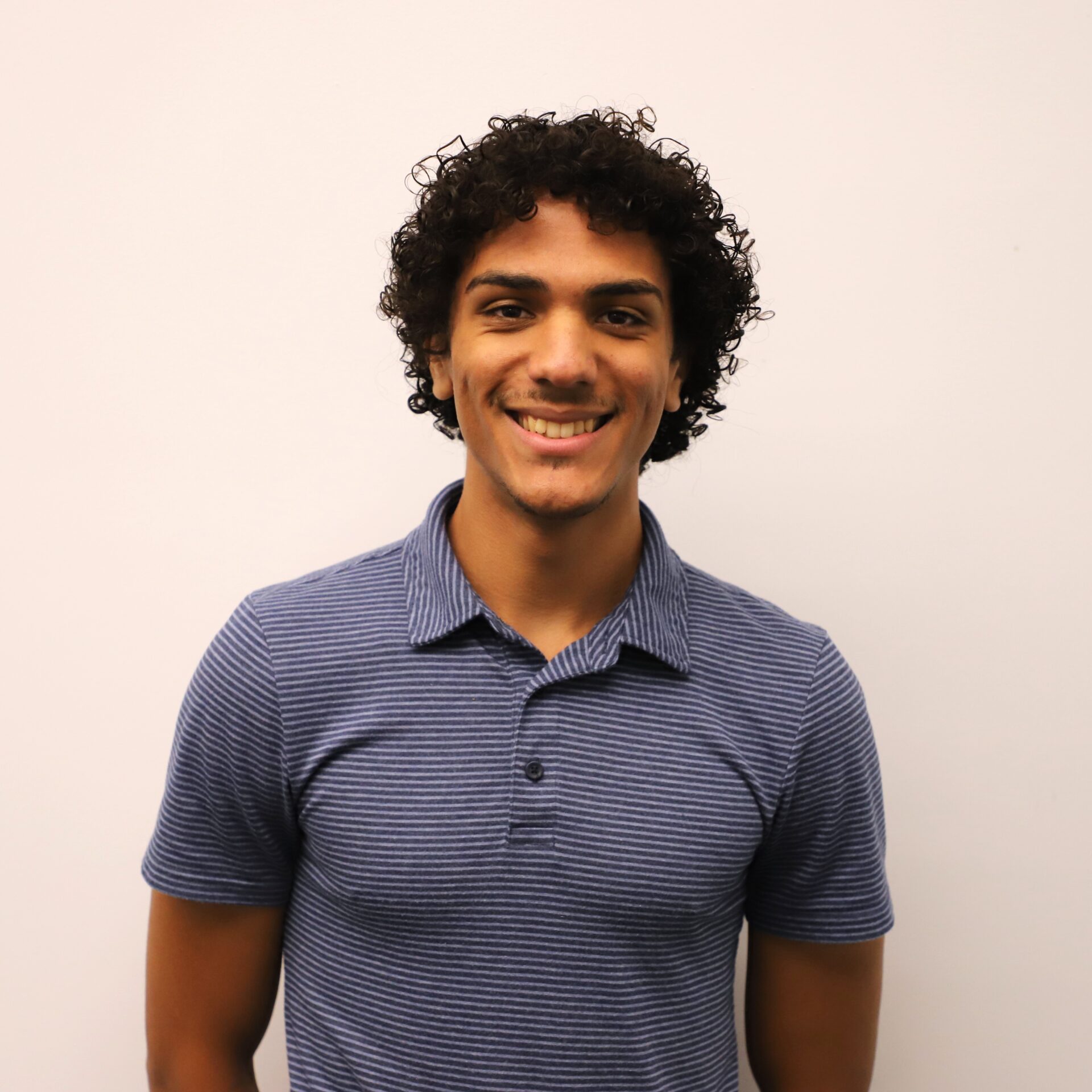 A young man with short curly hair, wearing a blue striped polo shirt, stands against a plain white background and smiles at the camera.