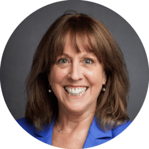 A woman with shoulder-length brown hair, wearing a blue blazer, smiling at the camera against a plain gray background.