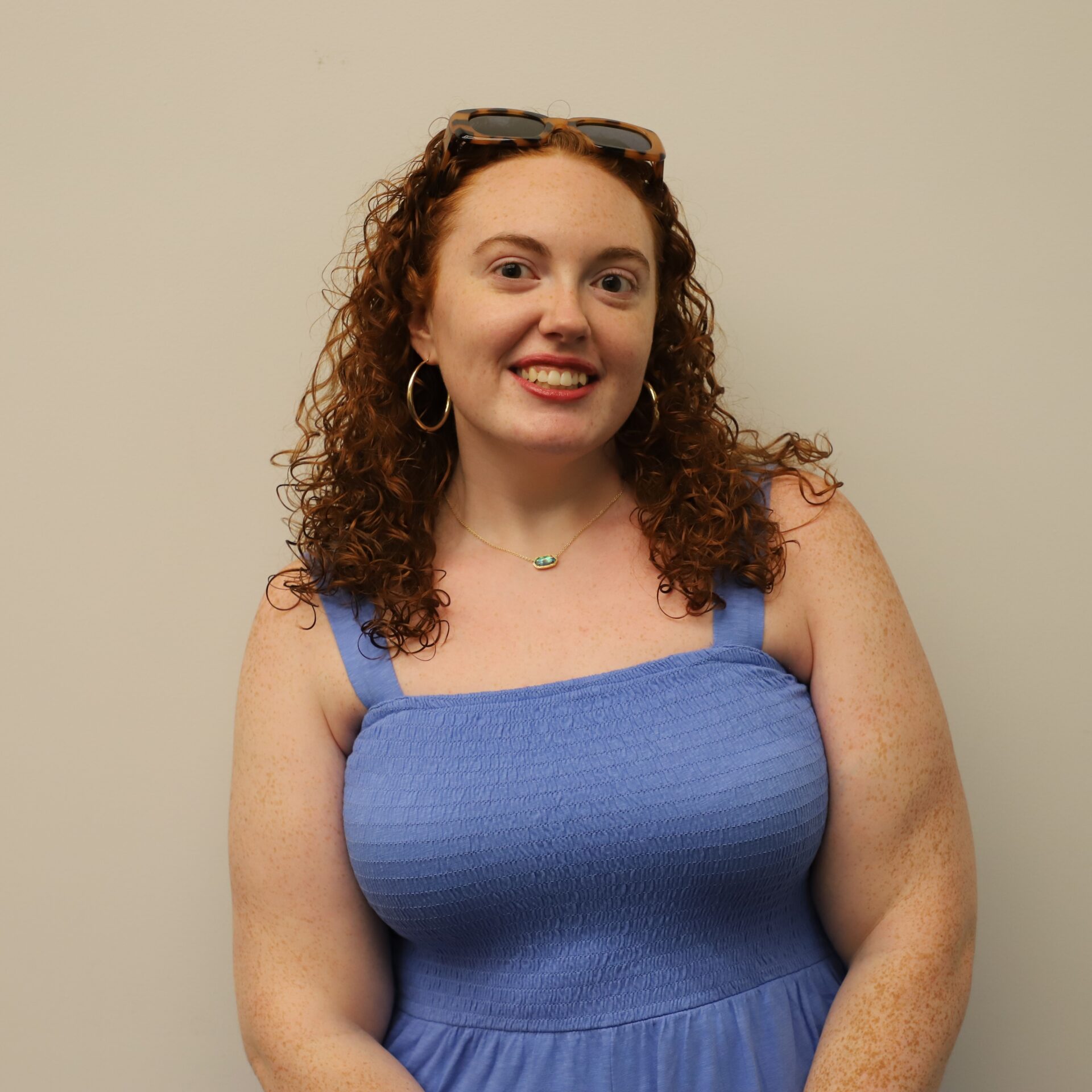 Woman with curly red hair wearing a blue sleeveless dress and sunglasses on her head, standing against a plain light-colored wall.