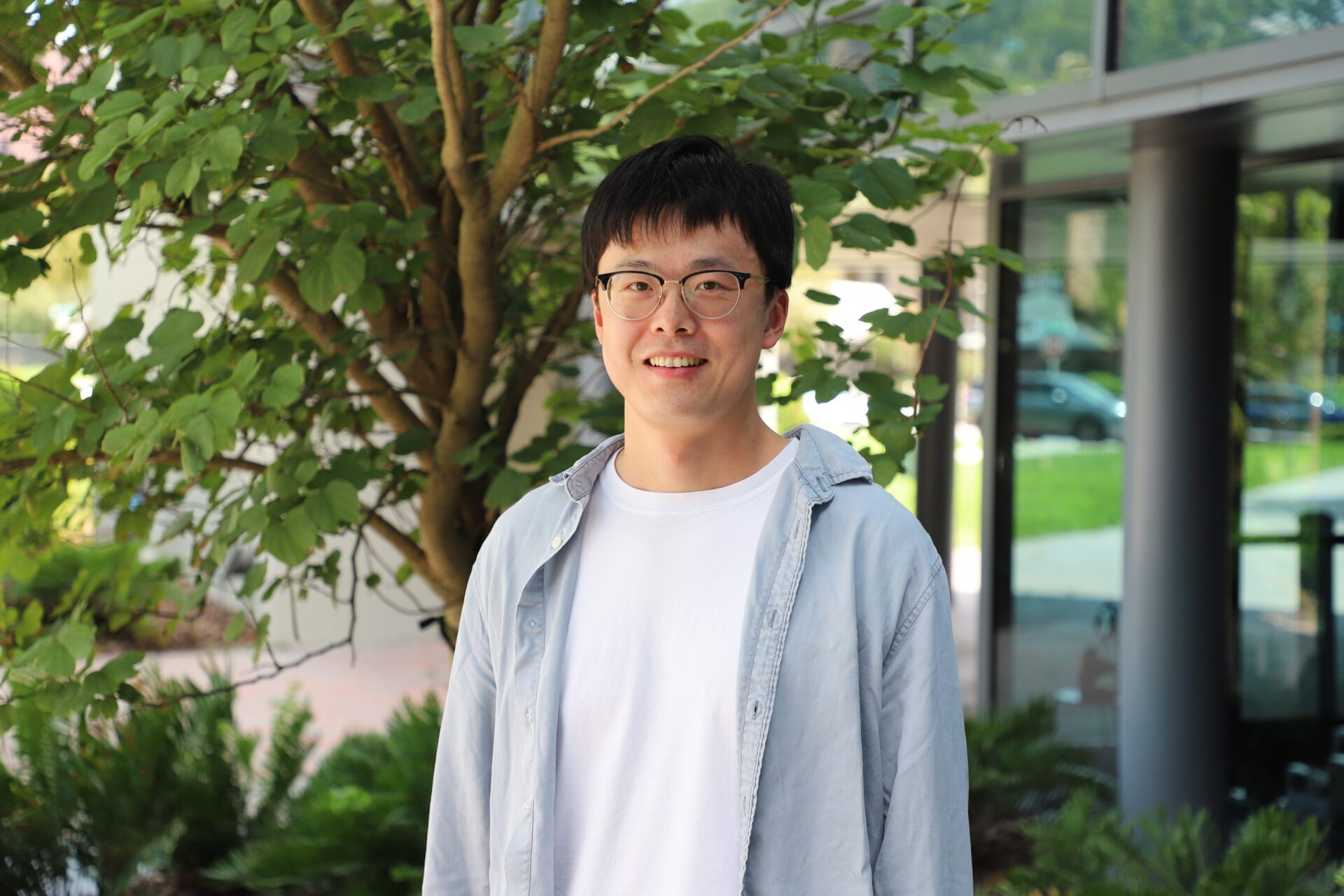 A man wearing glasses, a white t-shirt, and a light blue shirt stands outside in front of a tree and building, smiling at the camera.