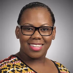 A woman with glasses and braided hair smiles at the camera. She is wearing a colorful patterned top against a neutral gray background.