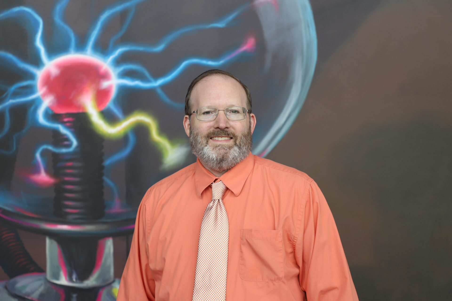 A man wearing glasses, an orange shirt, and a patterned tie stands in front of a mural depicting a colorful plasma ball.