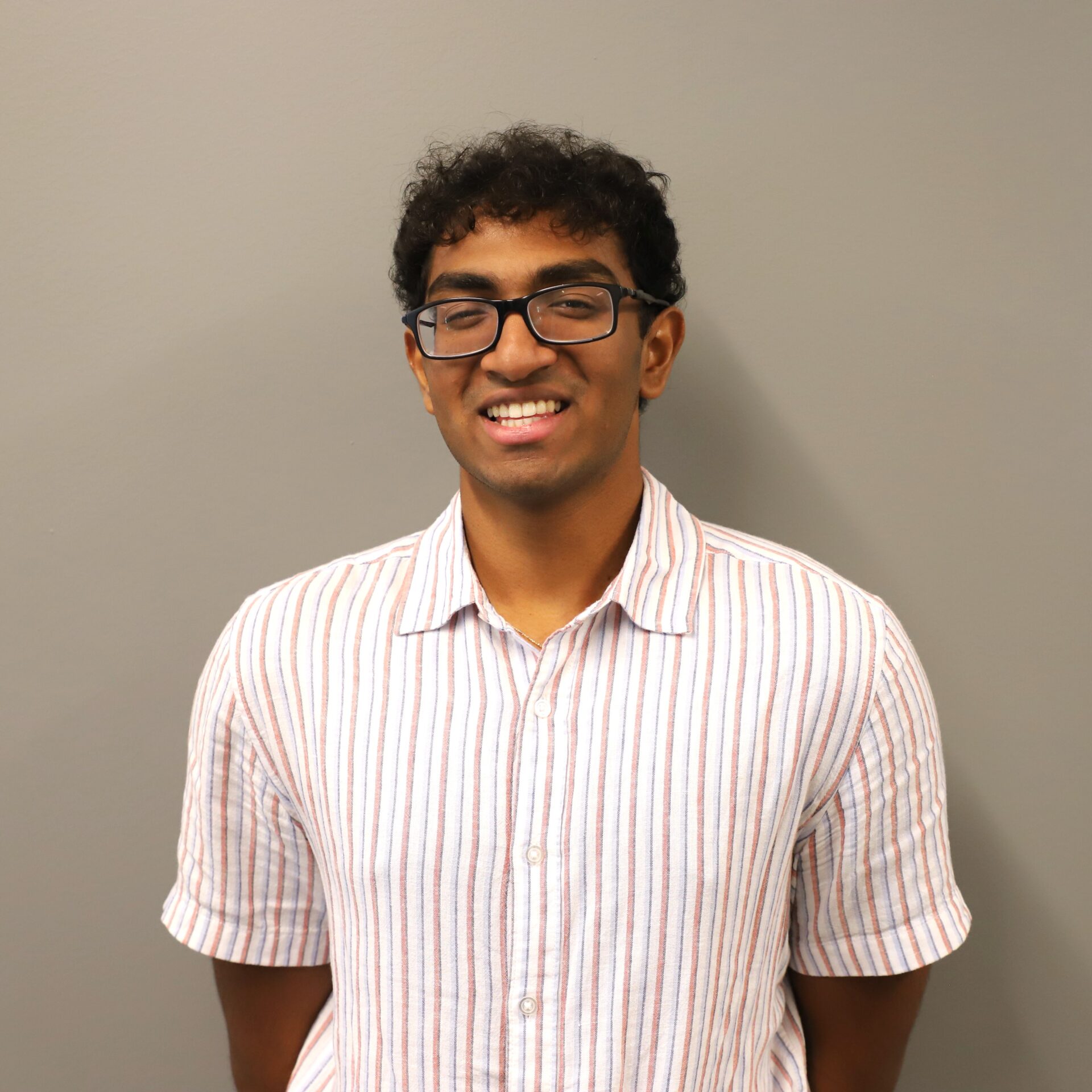 A person wearing glasses and a short-sleeve, white shirt with red and blue stripes stands in front of a plain gray wall, smiling at the camera.